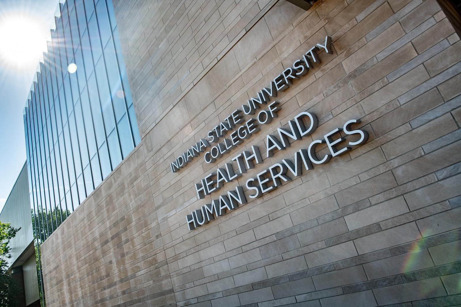 Exterior brick wall of Indiana State University College of Health and Human Services with large silver letters and a nearby glass building facade.