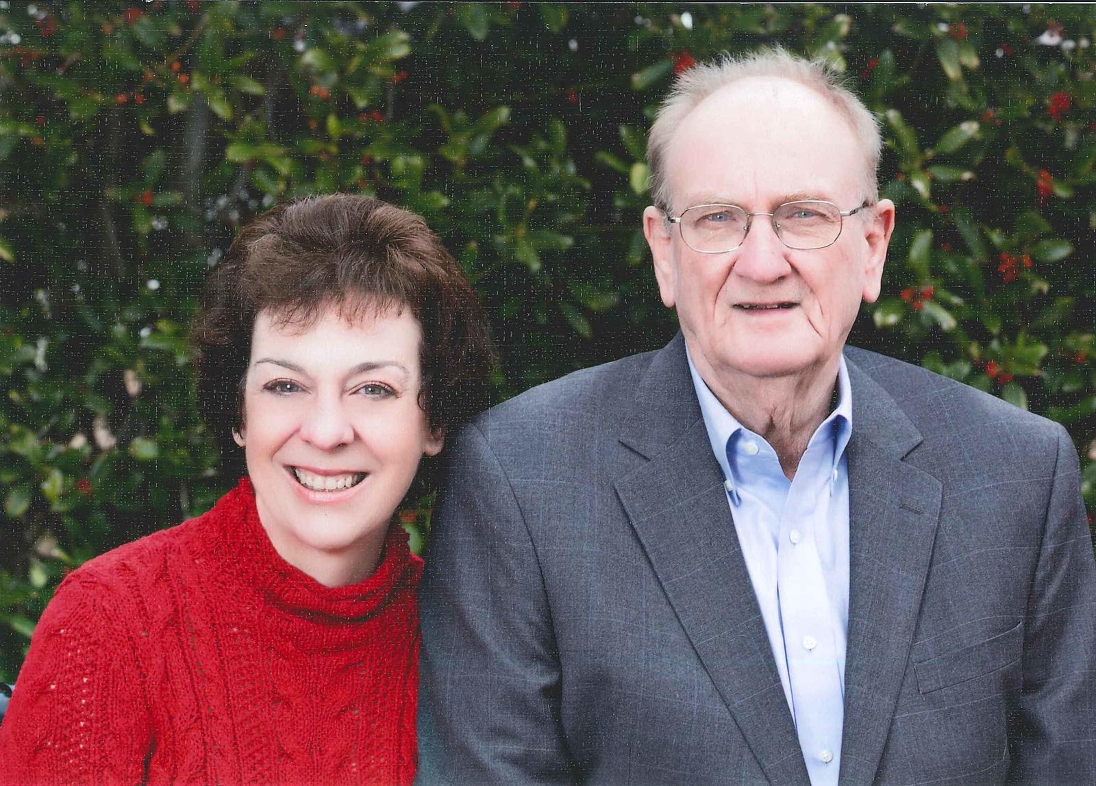 Smiling middle-aged woman in red sweater beside elderly man in gray suit and glasses, standing outdoors with green leafy background.