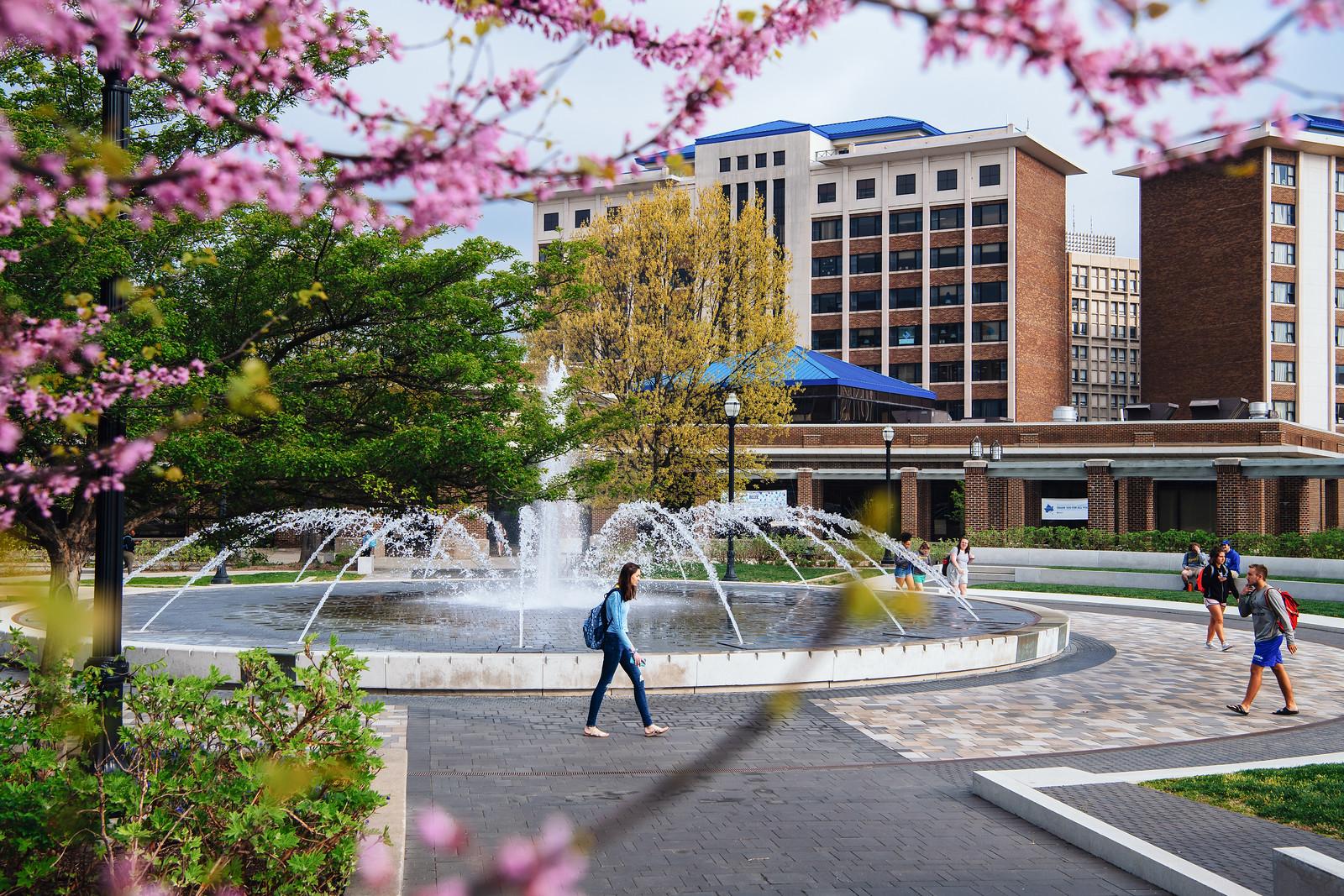 University campus scene with students walking near a large circular fountain, surrounding trees with spring blossoms, and multi-story buildings in the backgroun