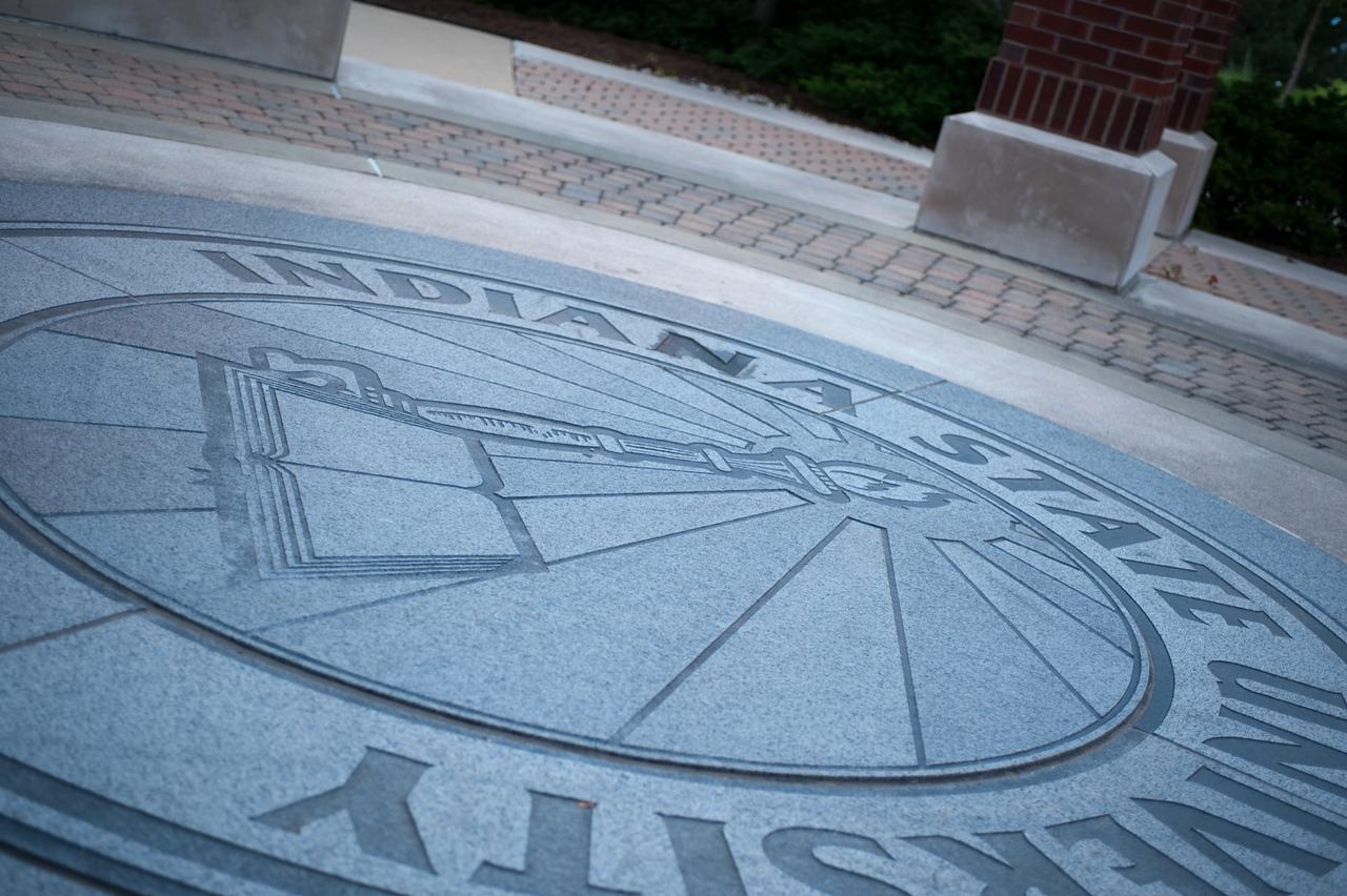 Engraved Indiana State University emblem on a stone surface with brick pillars and landscaping in the background.