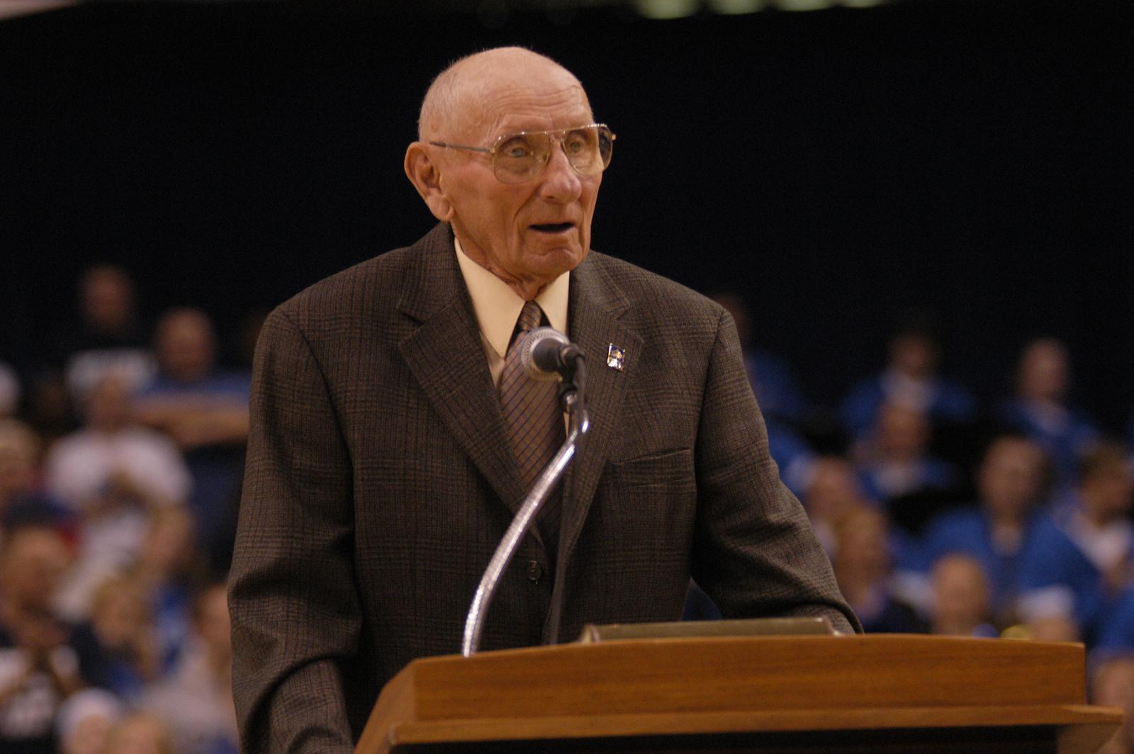 Elderly man in glasses and brown suit speaking at a podium with microphone, audience blurred in background.