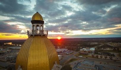 Golden dome of Indiana State Capitol building with a sunset and cloudy sky in the background, overlooking a small cityscape.