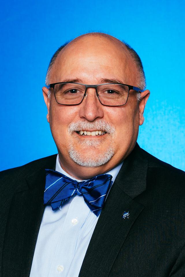 Smiling bald man with glasses and gray goatee wearing a black suit, blue dress shirt, and blue bow tie against a blue background.