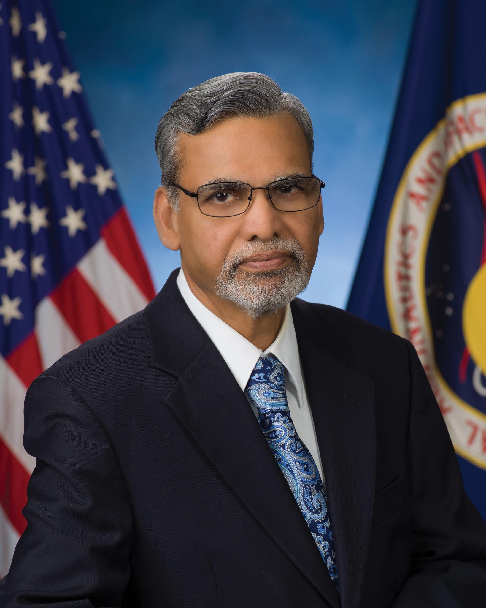 Middle-aged man with gray hair and beard wearing glasses, a dark suit, white shirt, and blue patterned tie, standing in front of American and NASA flags.