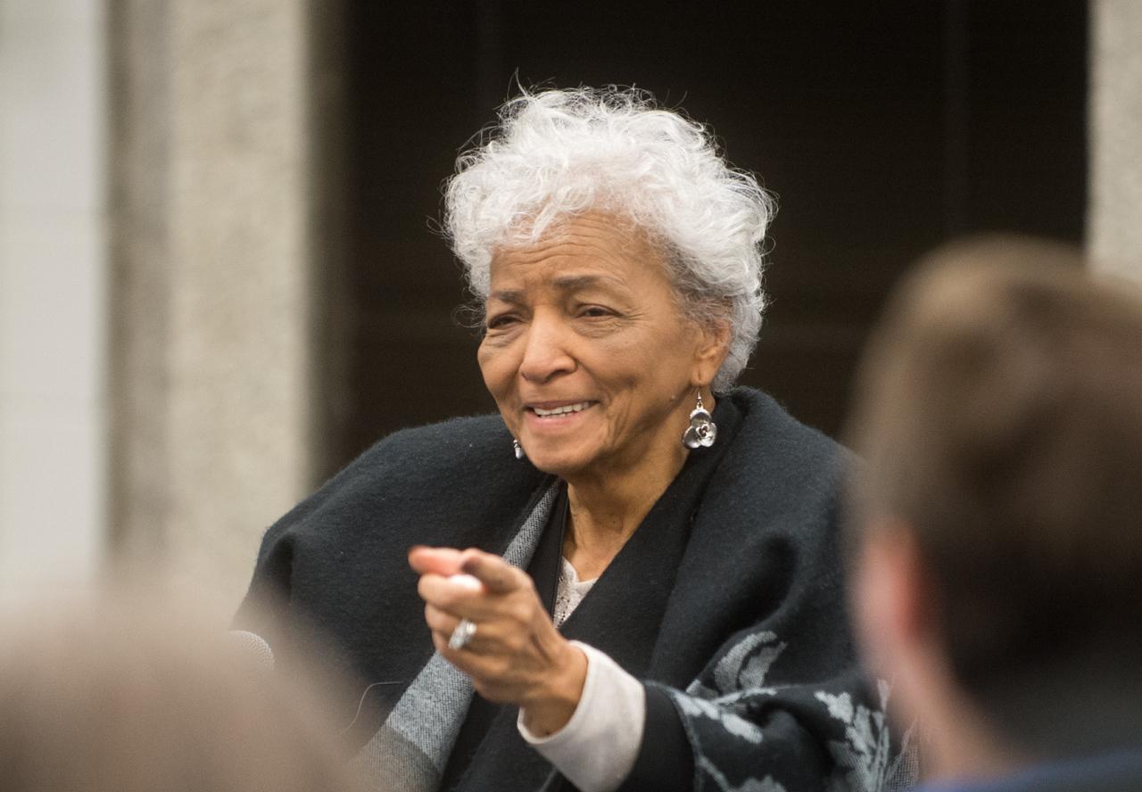 Elderly woman with curly white hair, wearing earrings and a dark shawl, smiling and pointing forward during an outdoor event.