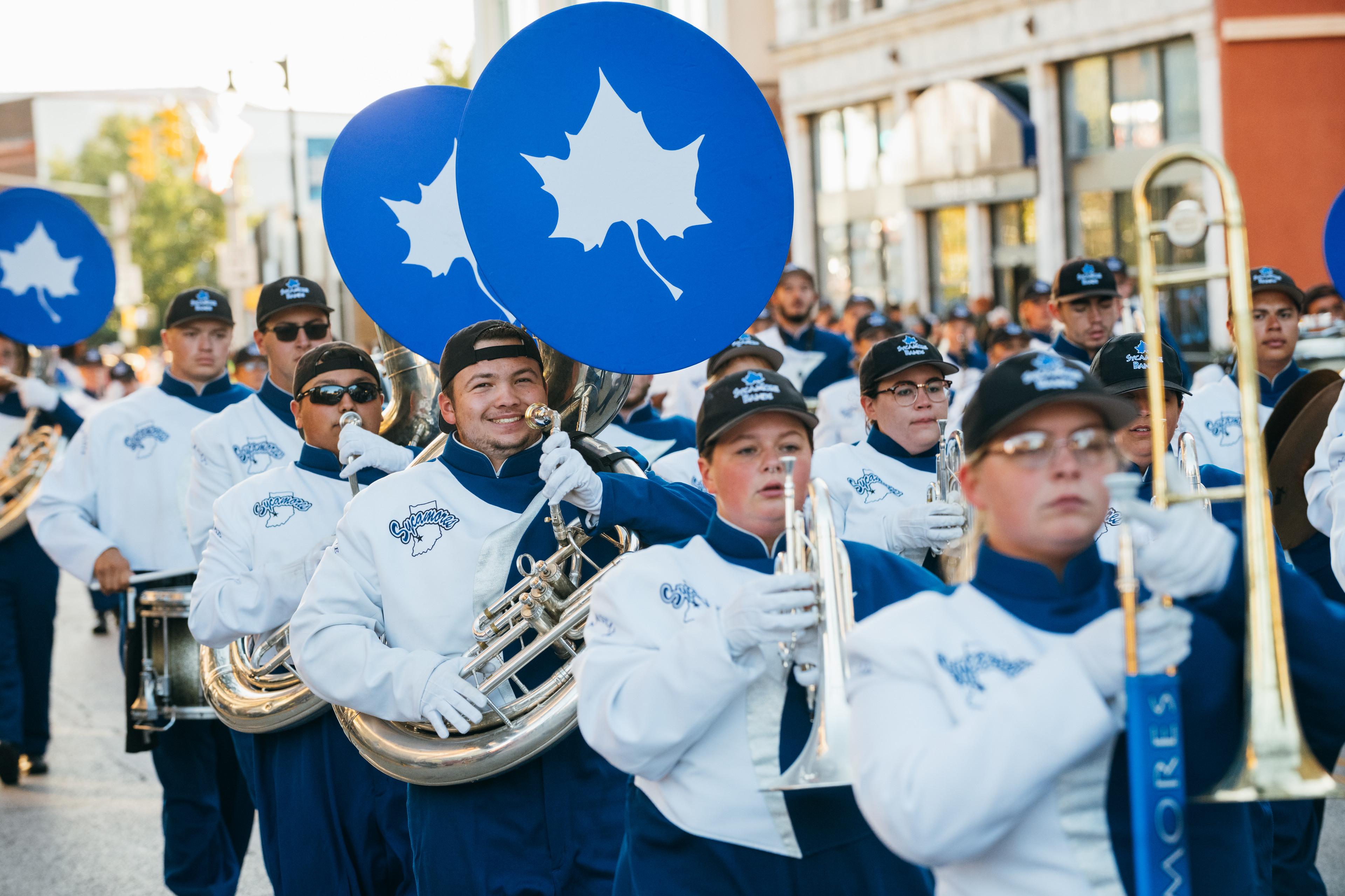 Marching band in blue and white uniforms plays brass and percussion instruments during a daytime parade on a city street.