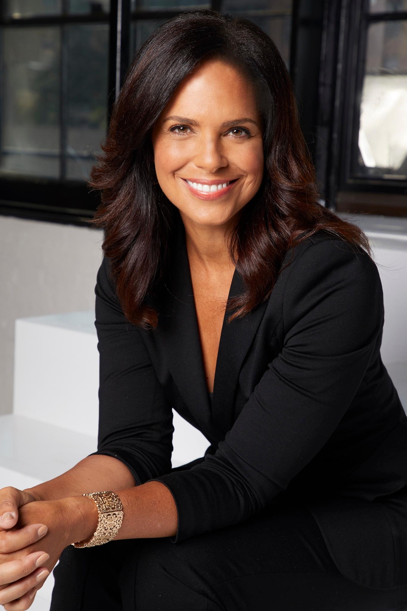 Smiling woman with medium-length dark hair, wearing a black blazer and pants, seated indoors with crossed hands and gold bracelet on wrist.