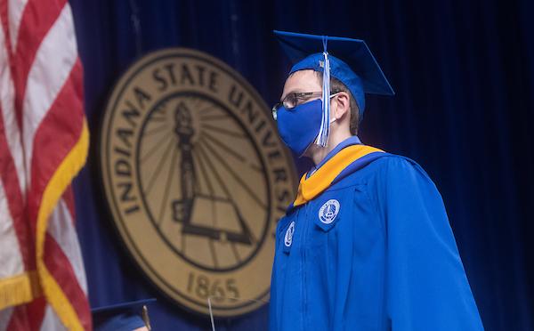 Graduation ceremony with a graduate in blue cap and gown standing near an Indiana State University seal and a U.S. flag.