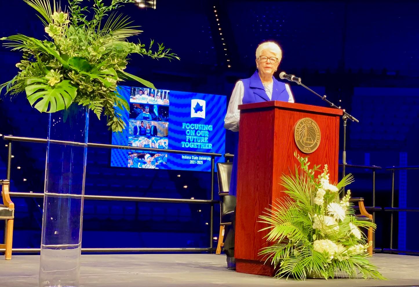 Woman in blue vest speaking at a wooden podium with Indiana State University seal, large green floral arrangement nearby, and a presentation screen in backgroun