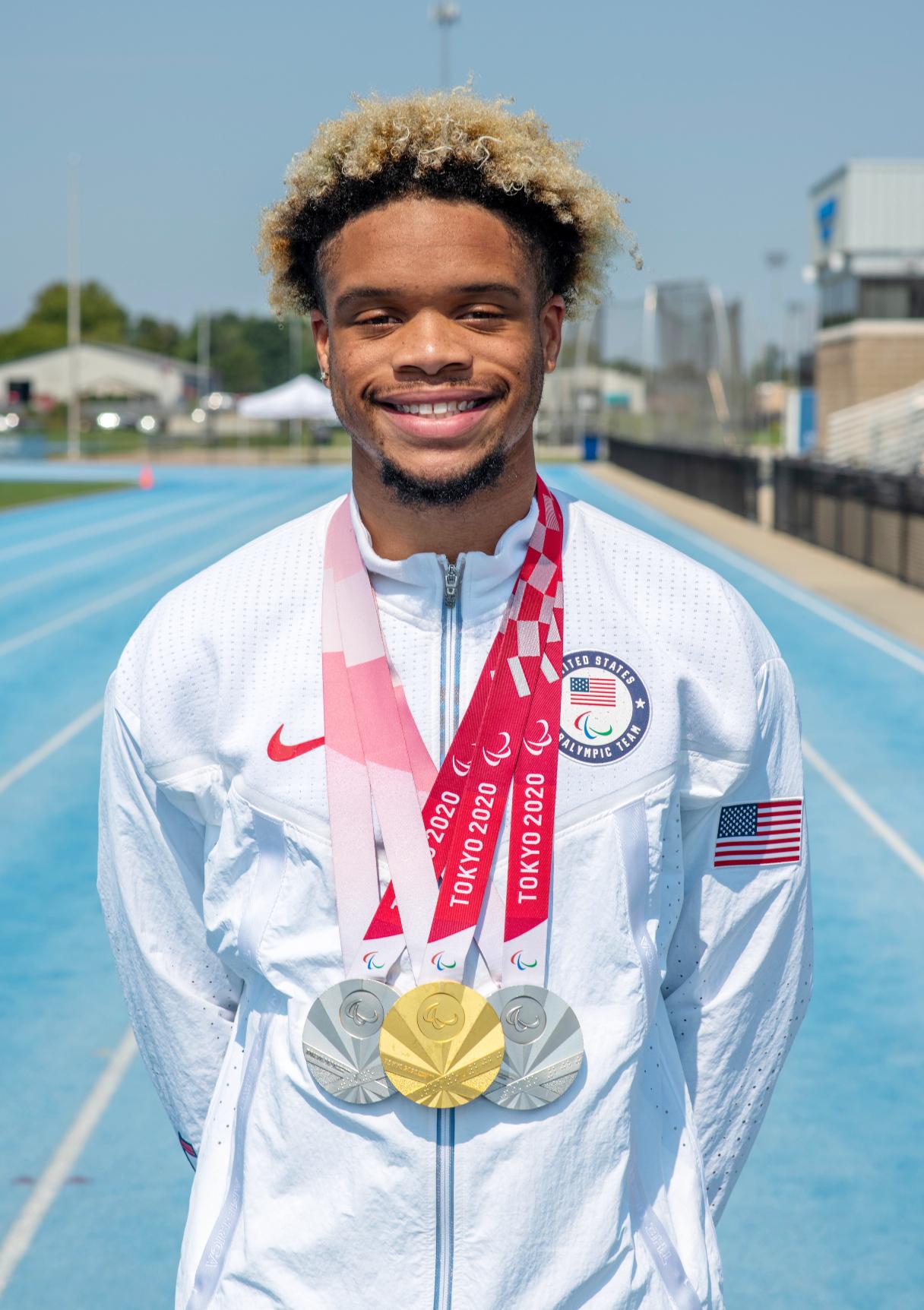 Young male athlete with blonde curly hair wearing a white US Paralympic jacket standing on a blue track with three Tokyo 2020 Paralympic medals around his neck.