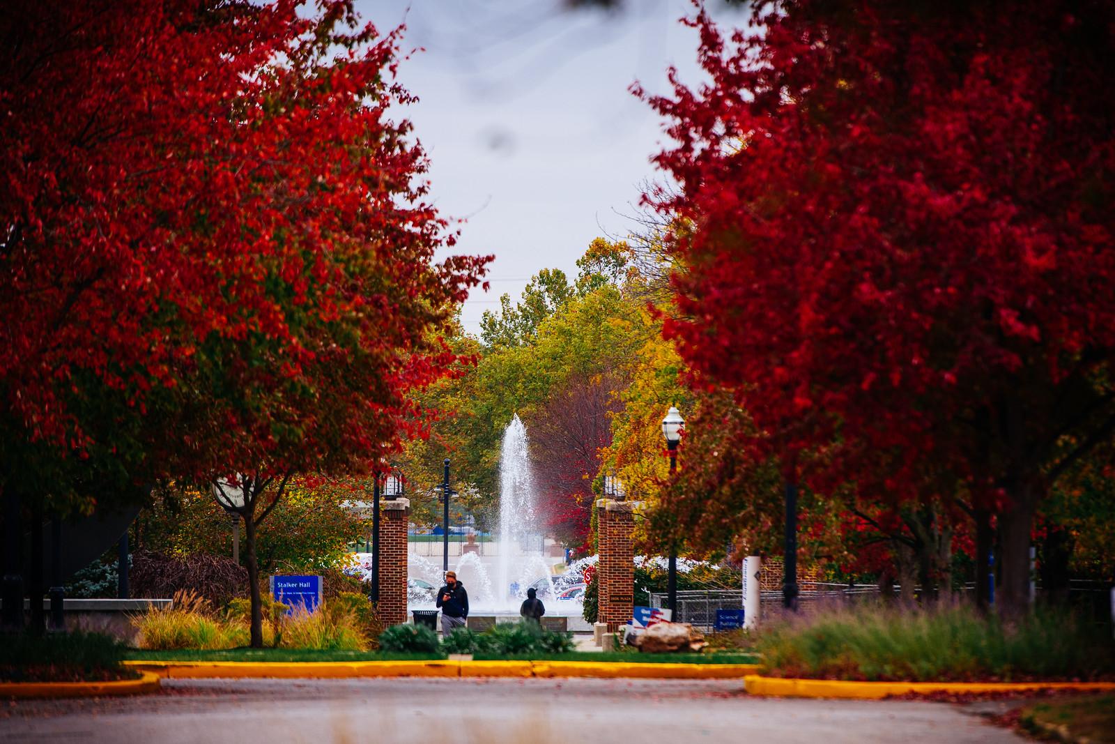 Fountain centered between red and green leafy trees lining a walkway, with two people walking near brick pillars on a cloudy day.