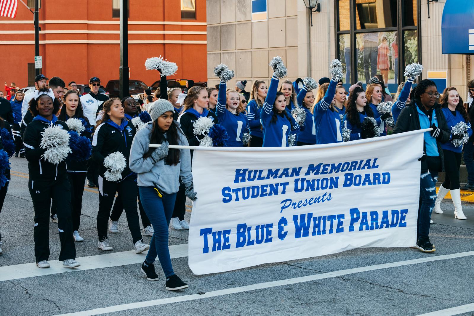 Group of young people marching in a parade holding pom-poms and a banner reading Hulman Memorial Student Union Board presents The Blue & White Parade.