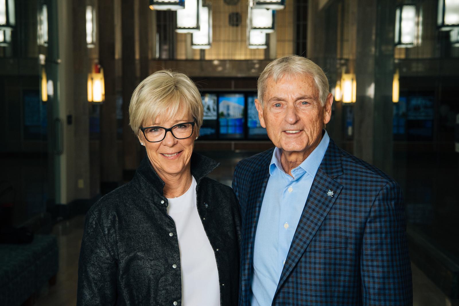 Older woman with short blonde hair and glasses wearing a black jacket next to an older man with gray hair wearing a blue checkered blazer and light blue shirt.