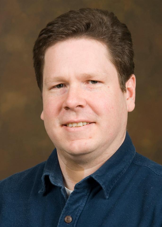 Middle-aged man with short brown hair wearing a dark blue collared shirt, smiling slightly against a brown background.