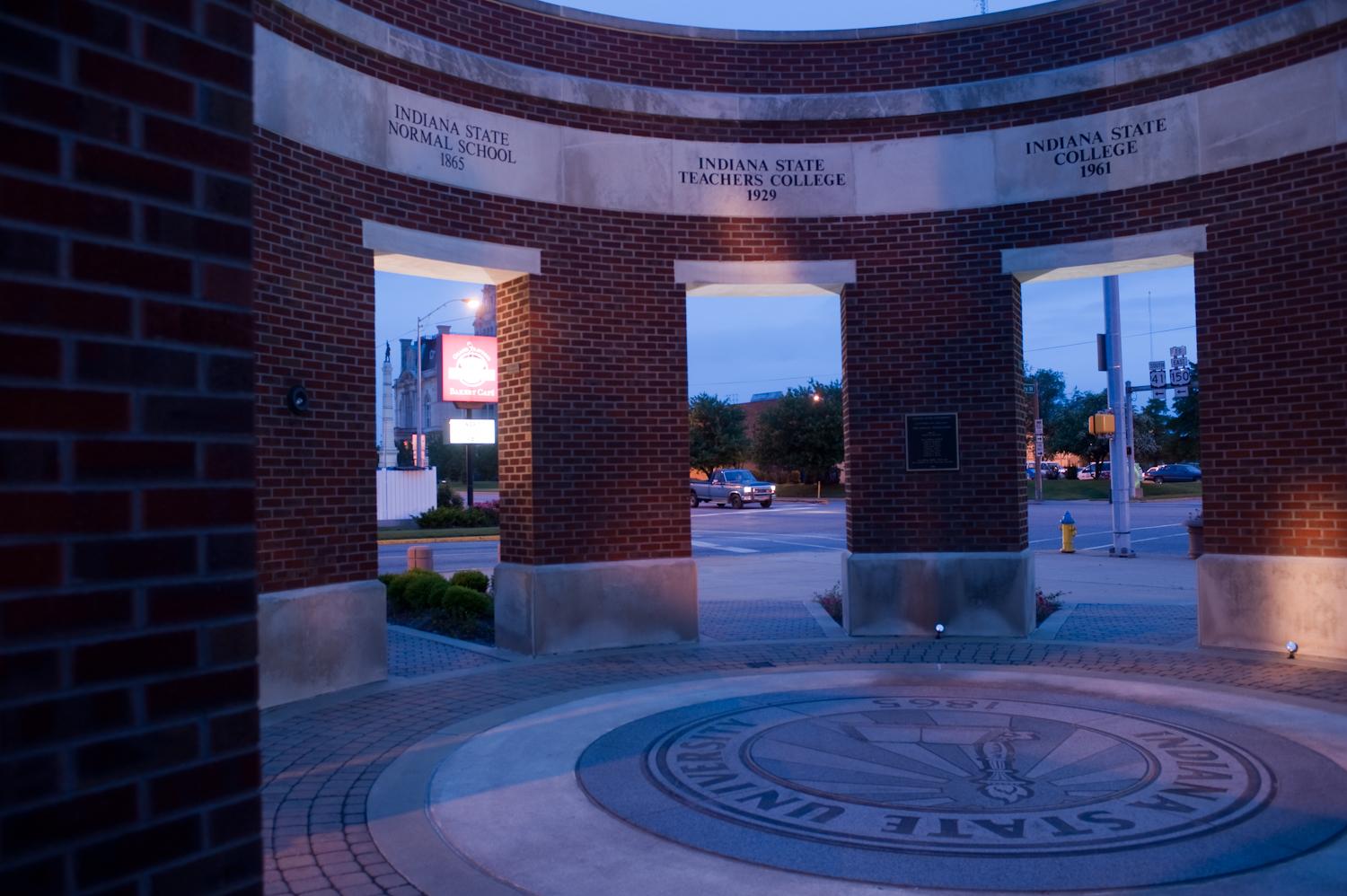 Brick archway at Indiana State University with inscriptions of the school's historical names and a circular seal on the ground at dusk.