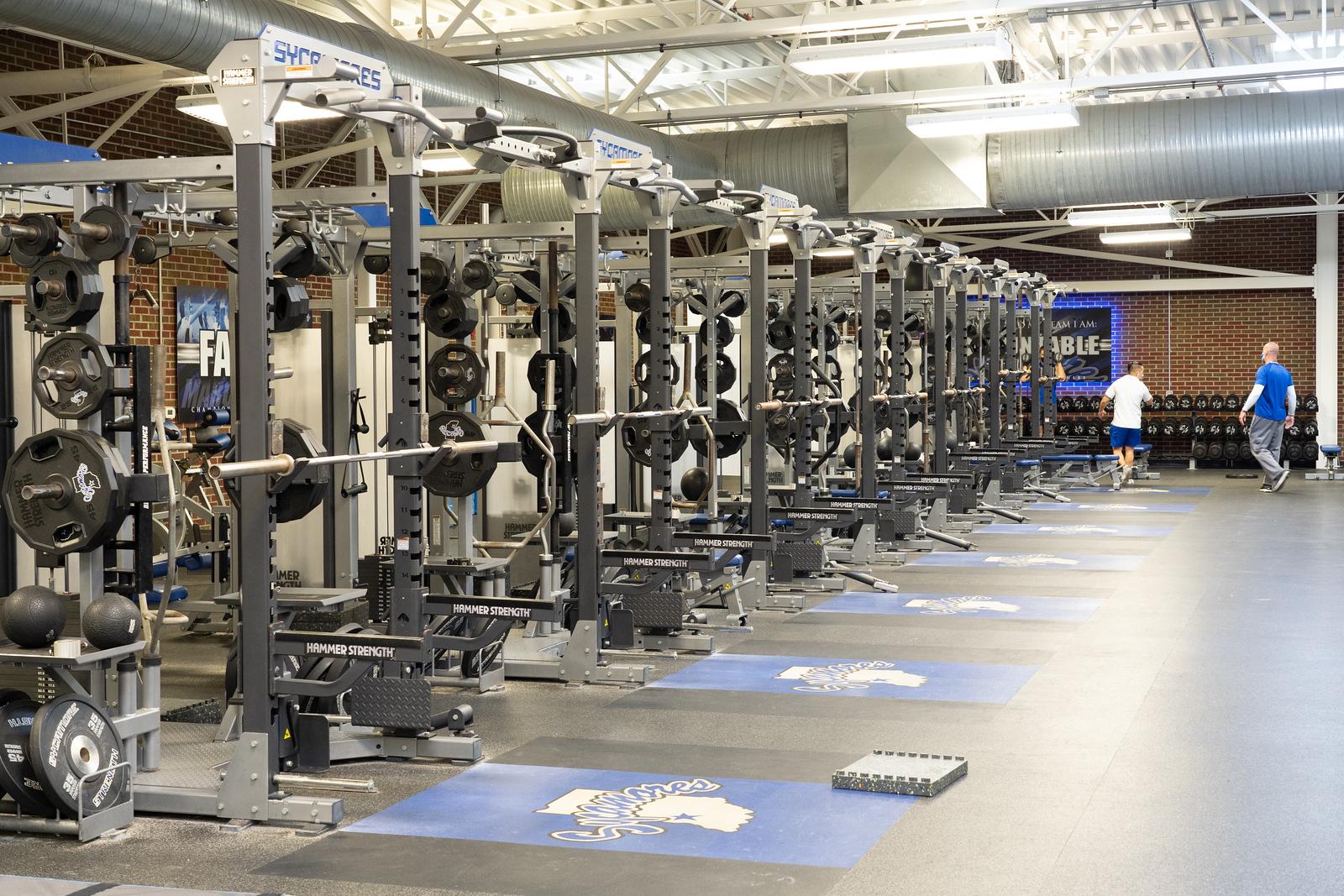 Row of weightlifting racks with bars and plates in an indoor gym, two people walking toward dumbbell area in the background, blue Sycamores logos on floor mats.