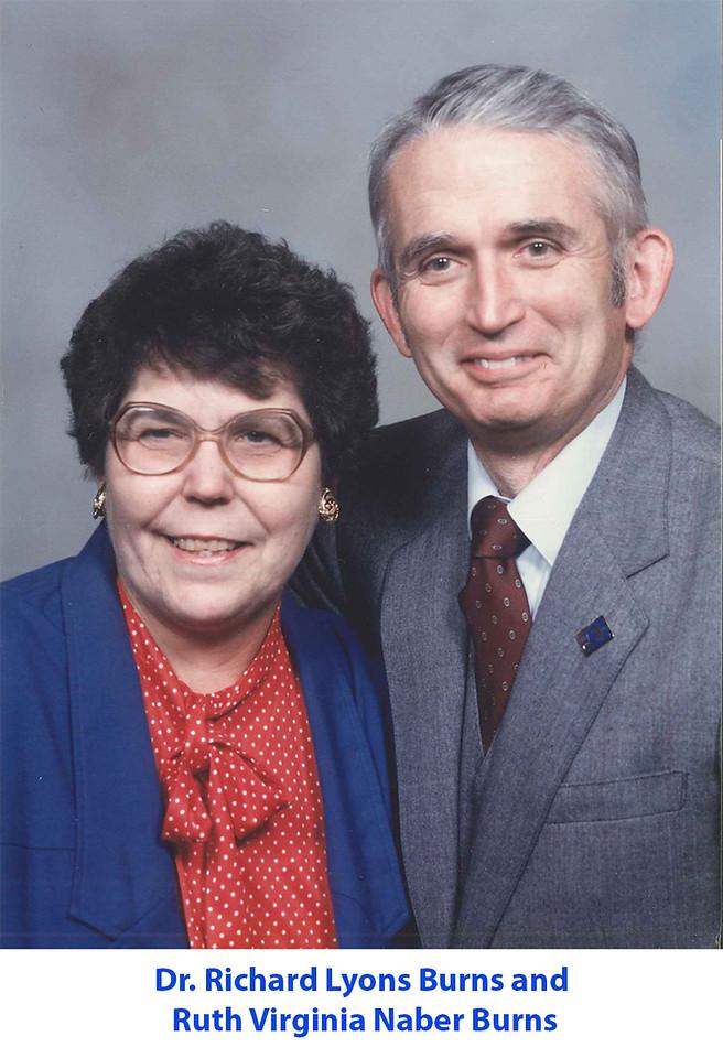 Middle-aged woman with glasses wearing a blue jacket and red polka-dot blouse, standing next to a smiling middle-aged man in a gray suit and tie.