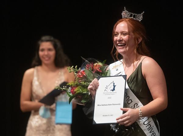 Red-haired woman smiling and wearing a crown and Miss Indiana State University sash, holding flowers and a certificate on a stage.