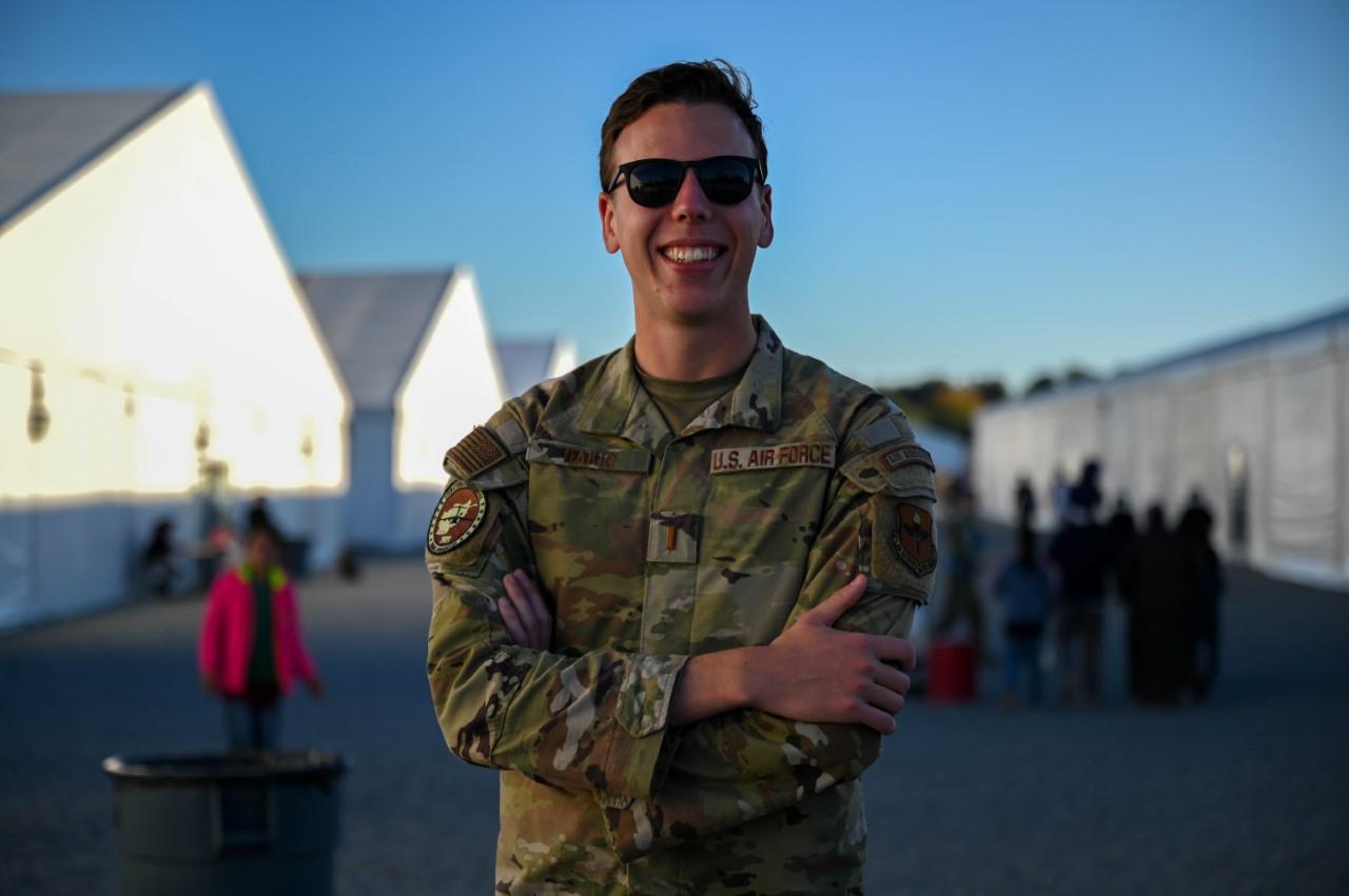 Smiling man wearing U.S. Air Force uniform and sunglasses standing outdoors with arms crossed, tents and people blurred in the background.