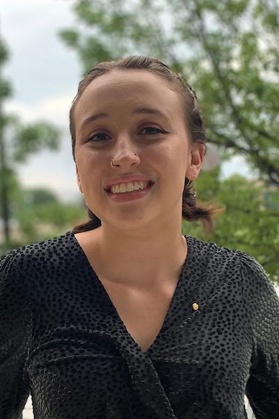 Young woman with brown hair in braided style, wearing black textured top with a small gold pin, standing outdoors with greenery and cloudy sky in the background