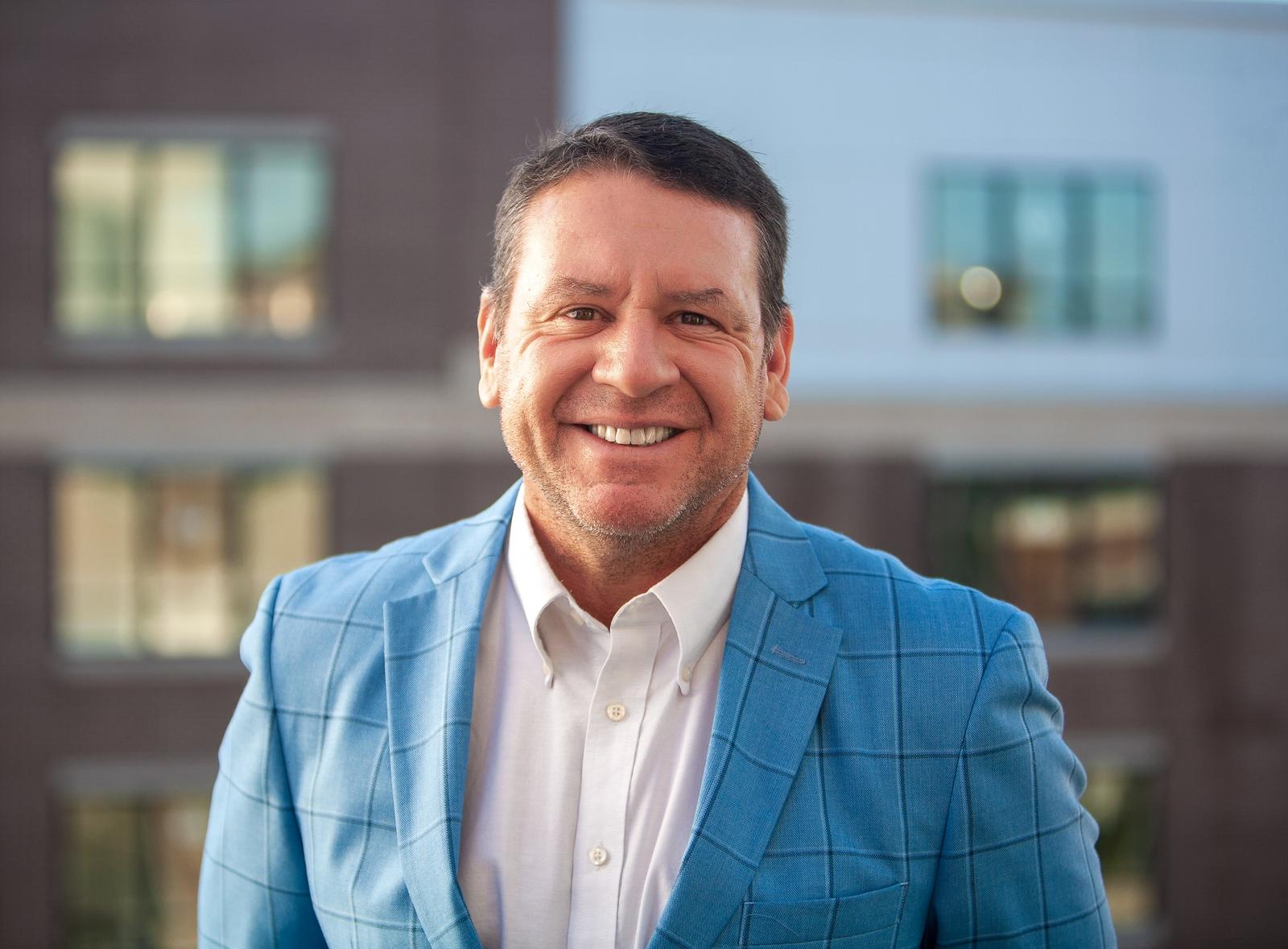 Middle-aged man wearing a blue checkered blazer and white shirt smiling with a blurred building background.