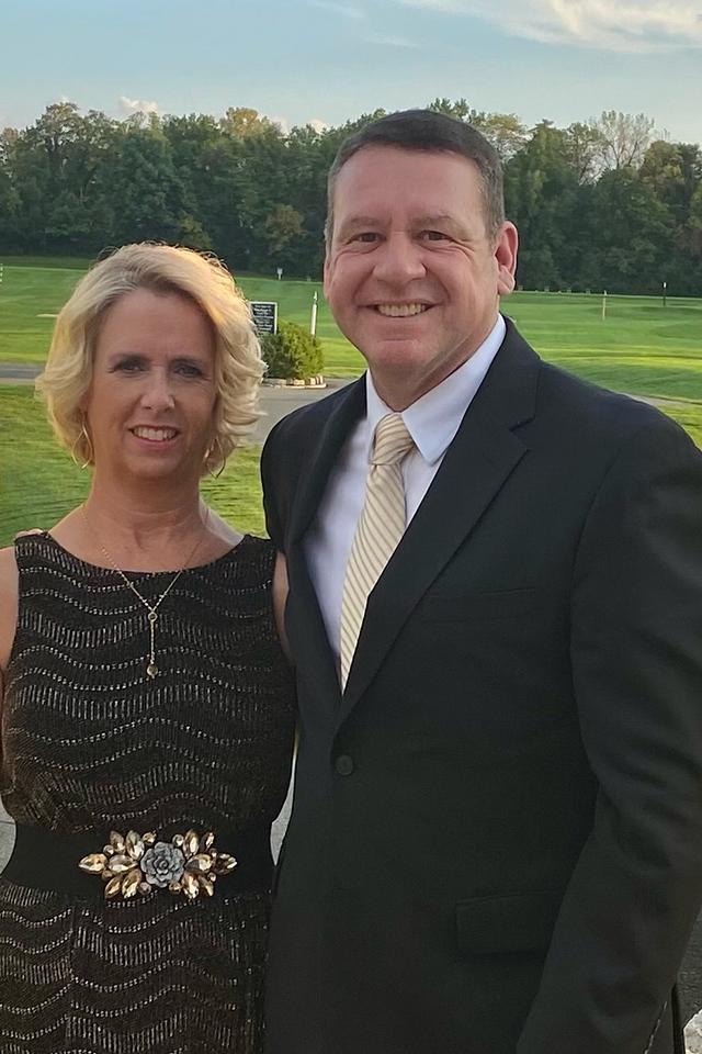 A man in a dark suit and light striped tie stands next to a woman in a black patterned dress with a decorative belt buckle, outdoors with grass and trees behind
