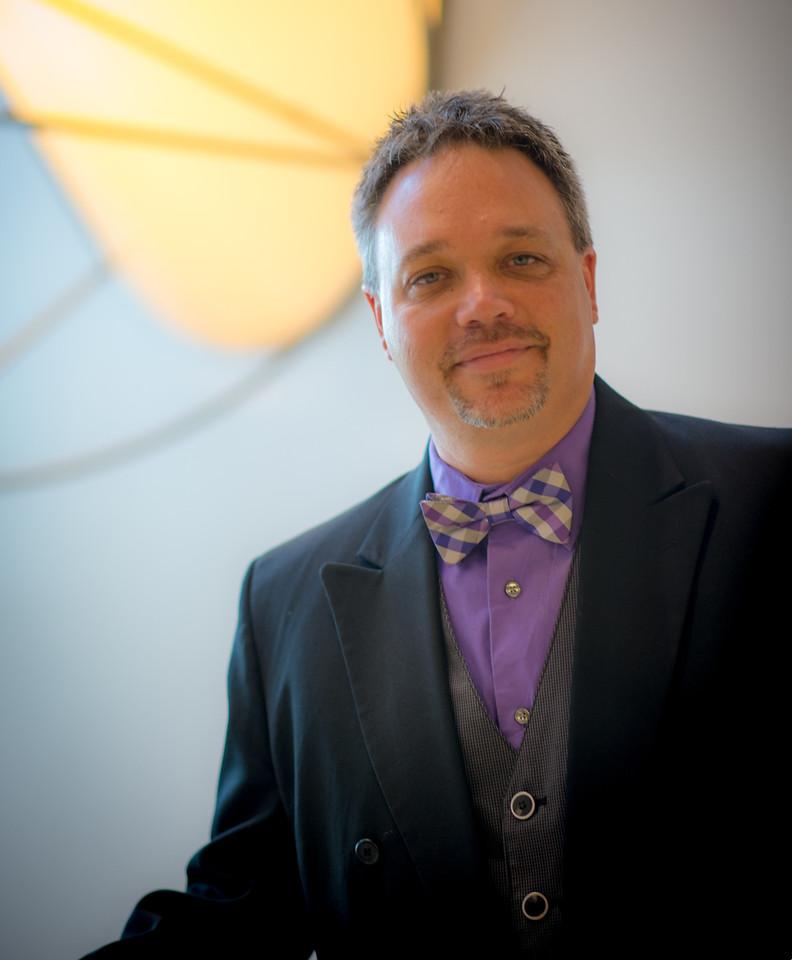 Man wearing a black jacket, purple shirt, patterned bow tie, and vest standing indoors with a large yellow geometric design in the background.
