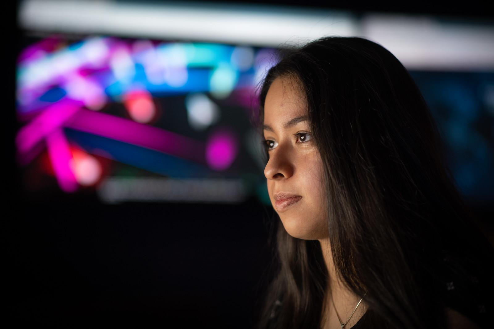 Young woman with long dark hair looking at a brightly colored digital screen in a dark room, focused and thoughtful.
