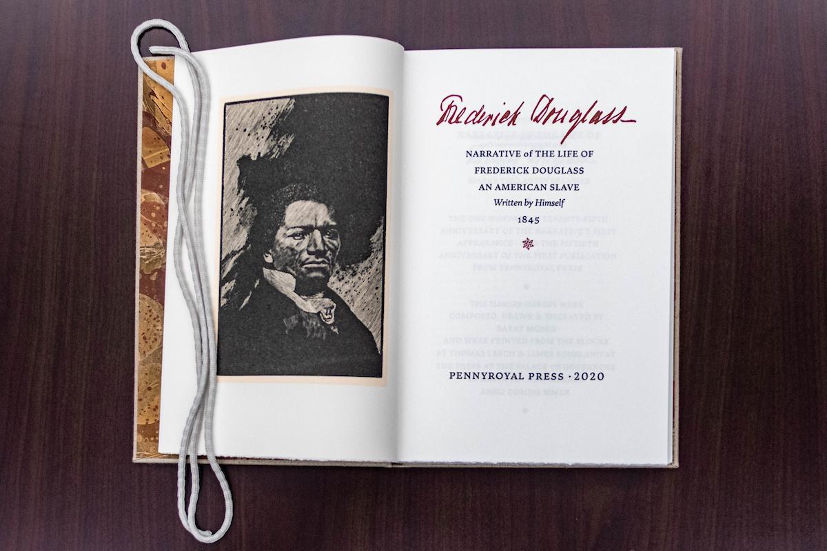 Open book showing a black and white portrait of Frederick Douglass on the left page and title page of his 1845 autobiography on the right page.