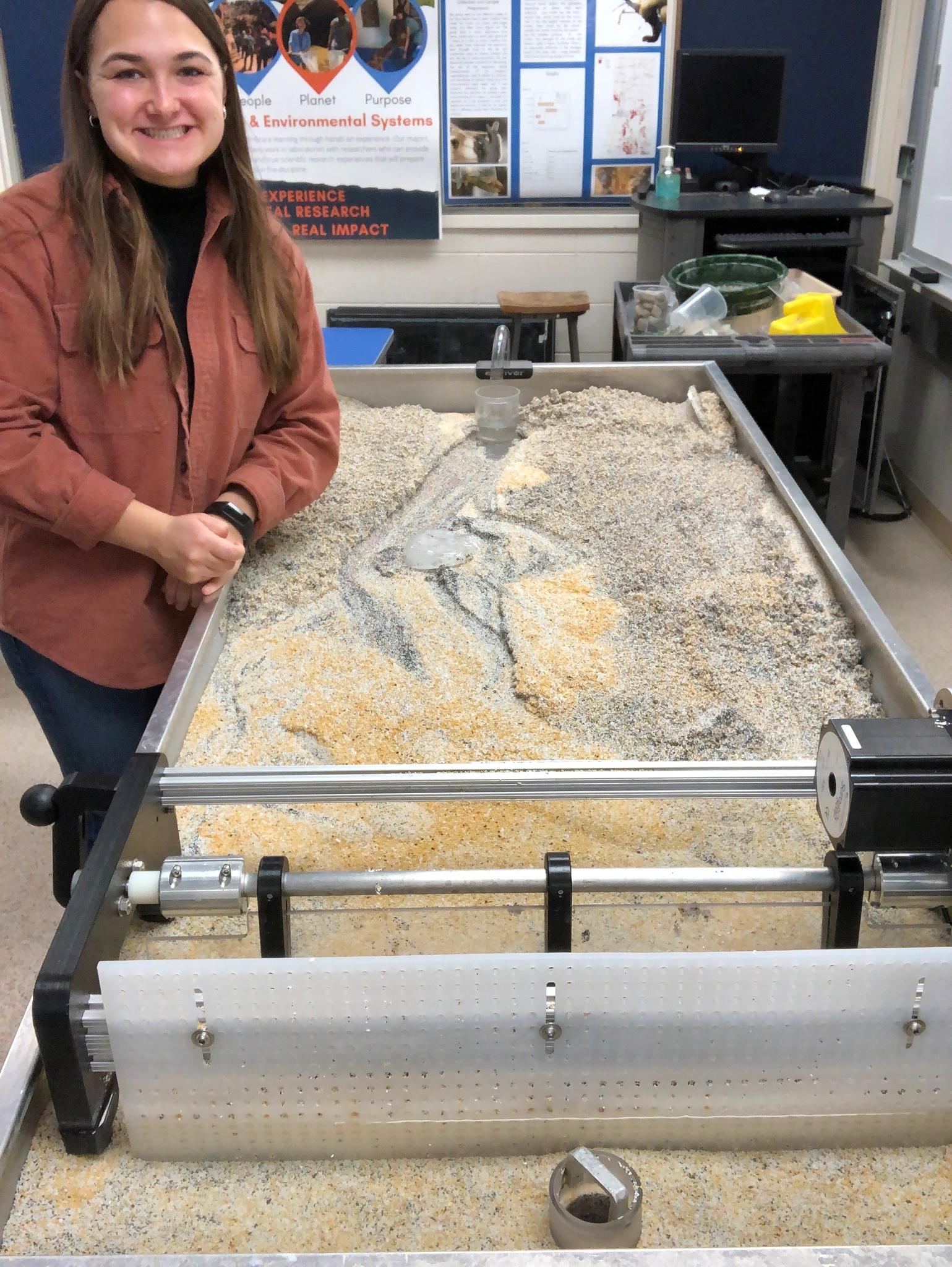 Young woman smiling beside a large sand and gravel table with a mechanized arm in a classroom with educational posters.