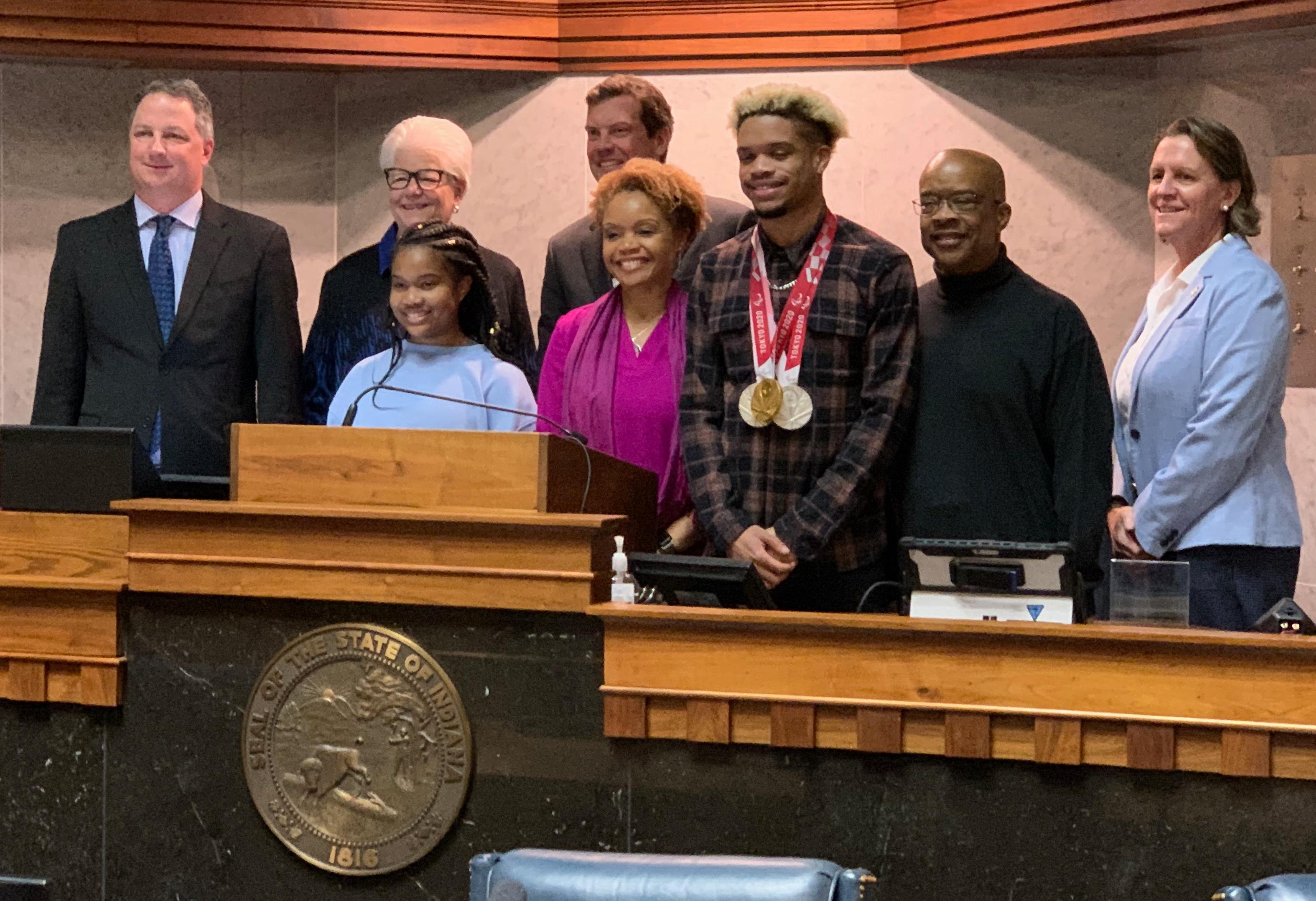 Seven people stand behind a wooden podium in a government chamber, one wearing Tokyo Paralympics medals around his neck. The Indiana state seal is visible.