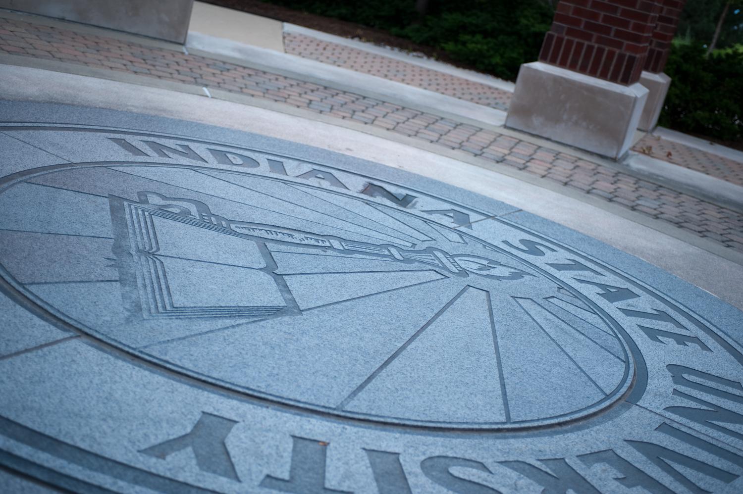 Indiana State University circular stone emblem set in ground near brick columns and paved walkway outdoors.