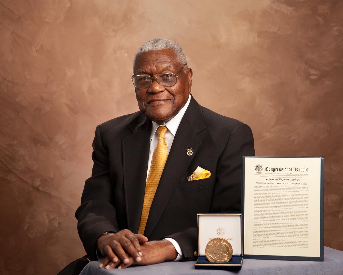 Elderly man in dark suit with gold tie and pocket square sits at table with a large medal in a case and a framed Congressional Record document.