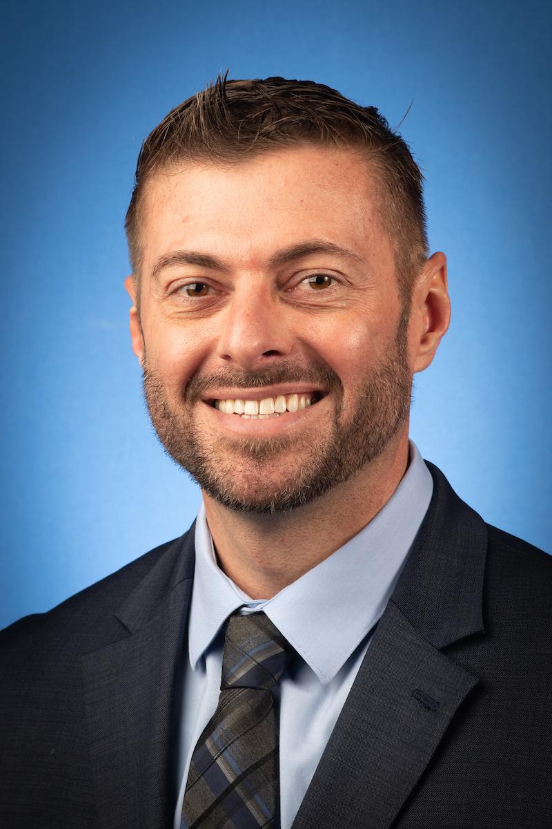 Light-skinned man with short brown hair and beard wearing a dark suit, light blue shirt, and patterned tie, smiling against a blue background.