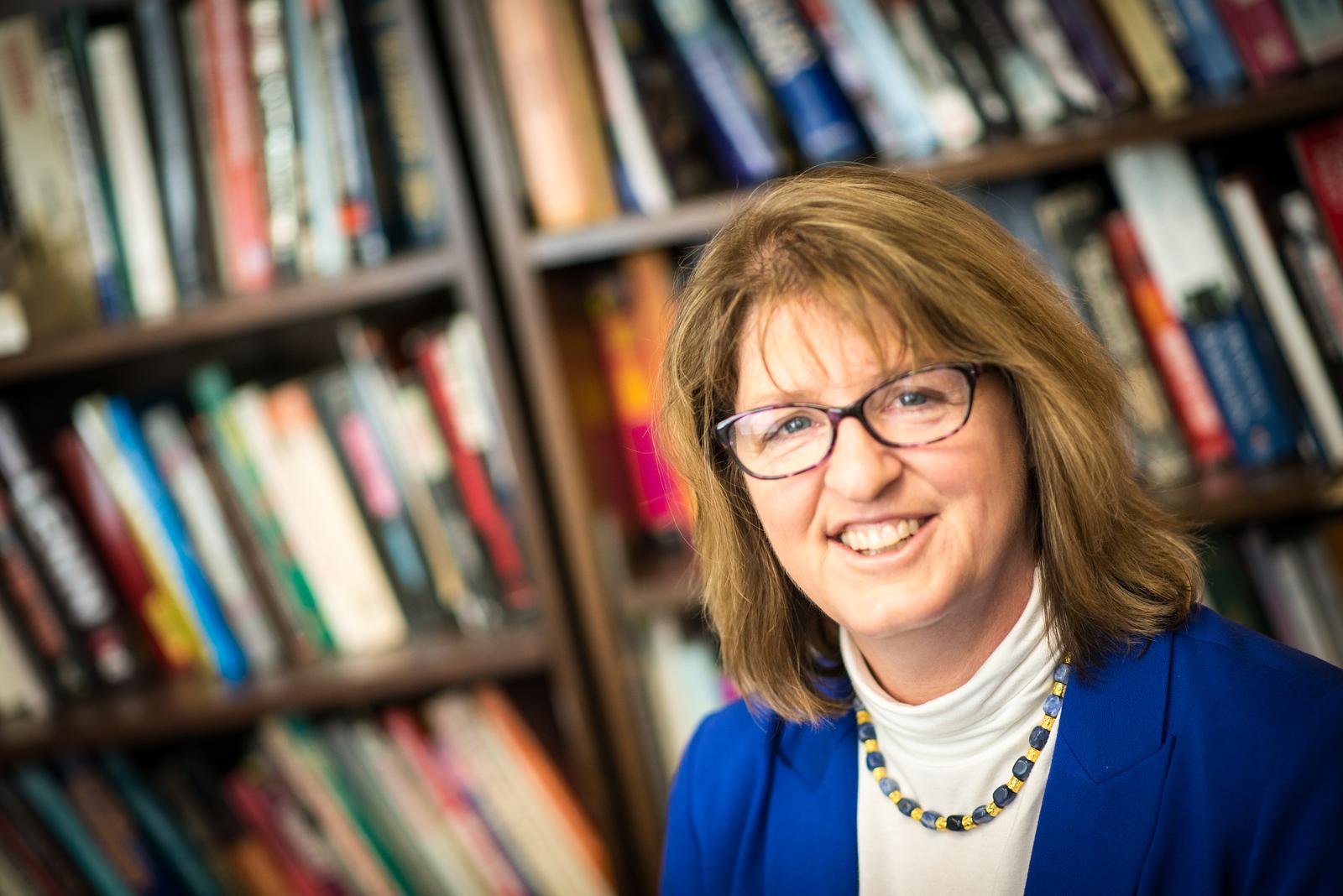 Woman with glasses, light brown hair, wearing a blue blazer and white turtleneck, smiling in front of a bookshelf filled with books.
