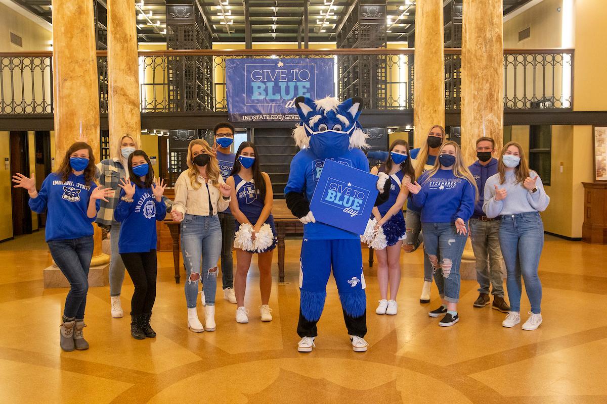 Group of people including cheerleaders and a mascot wearing blue, standing in a hall with a Give to Blue Day banner behind them.