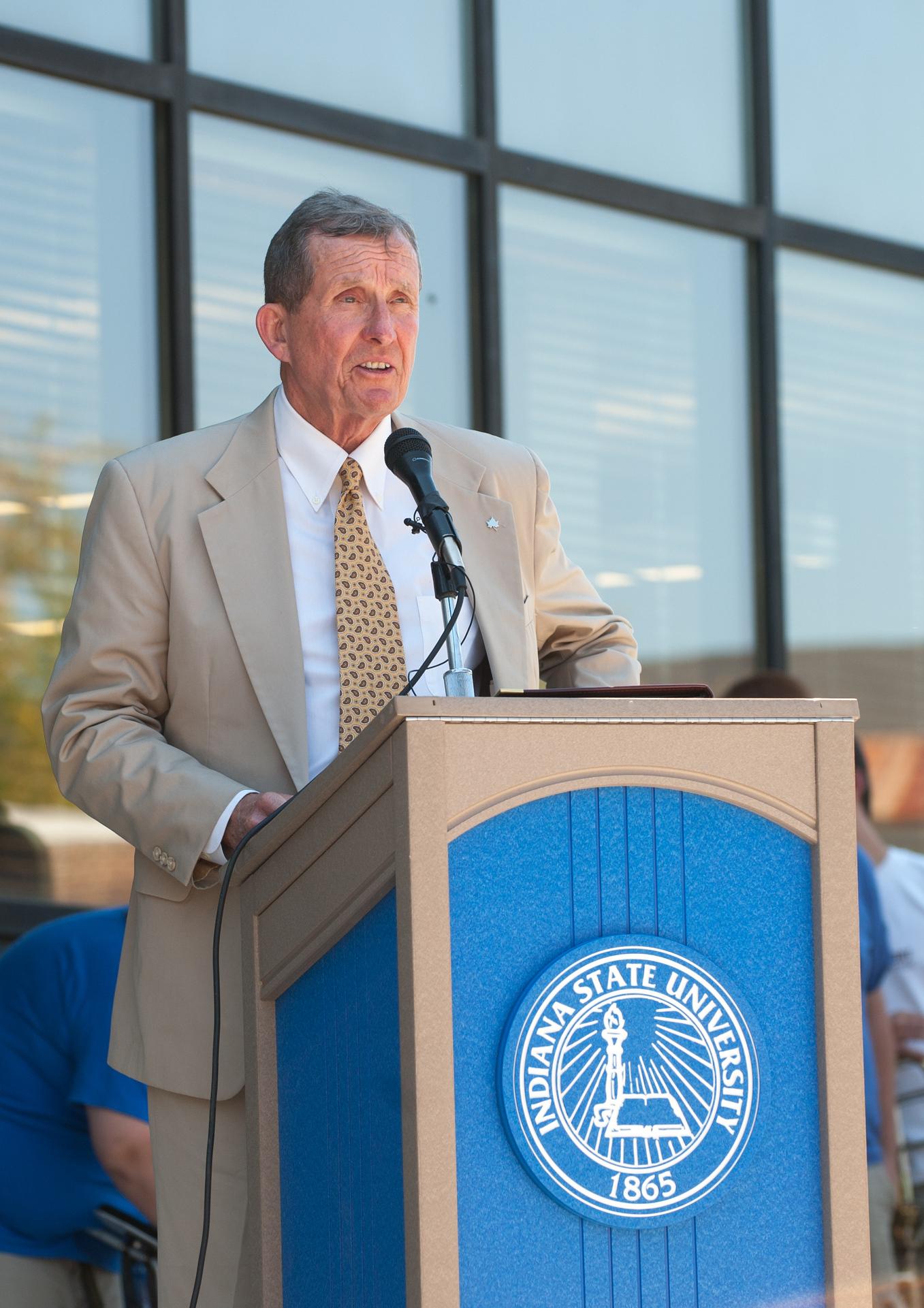 Man in beige suit and patterned tie standing behind a podium with Indiana State University seal, speaking into a microphone outdoors.