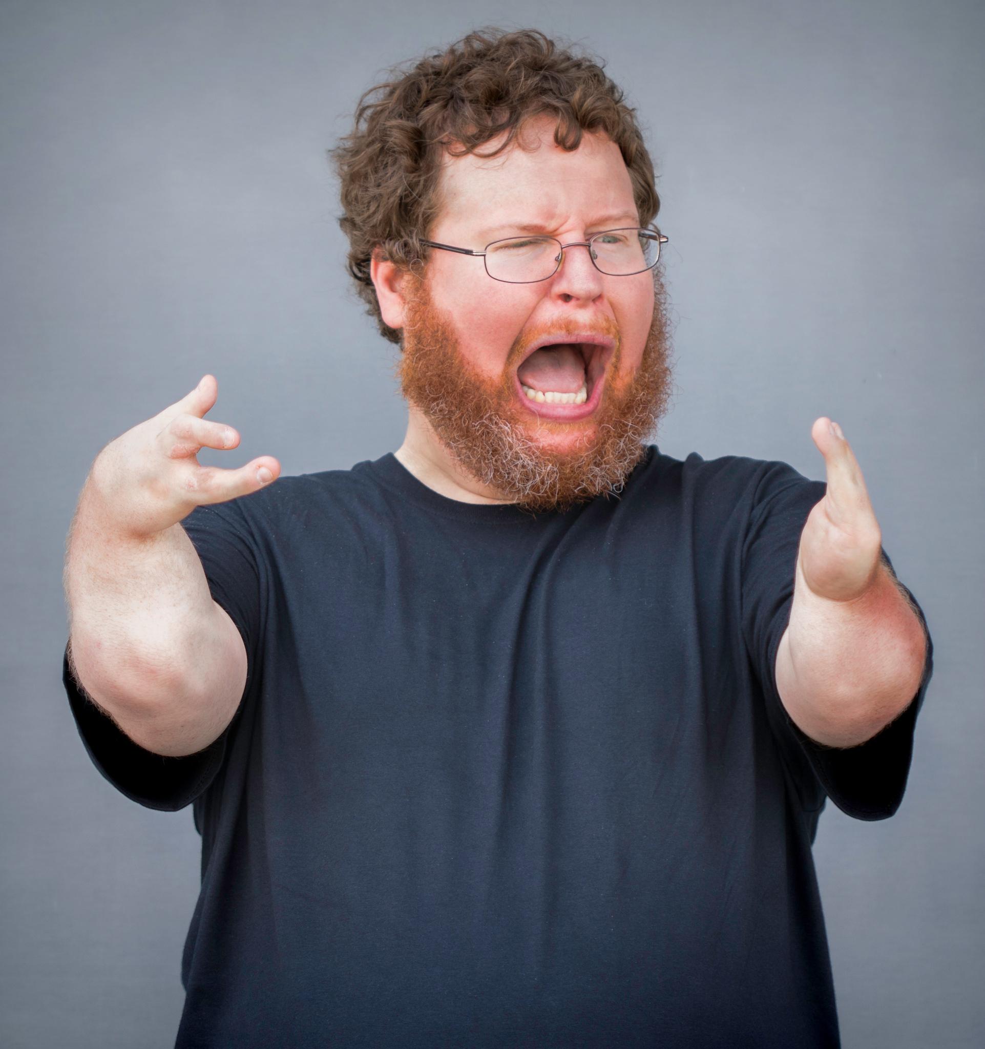 Man with red curly hair and beard wearing glasses and a black shirt, arms raised and mouth open as if shouting or singing against a plain gray background.