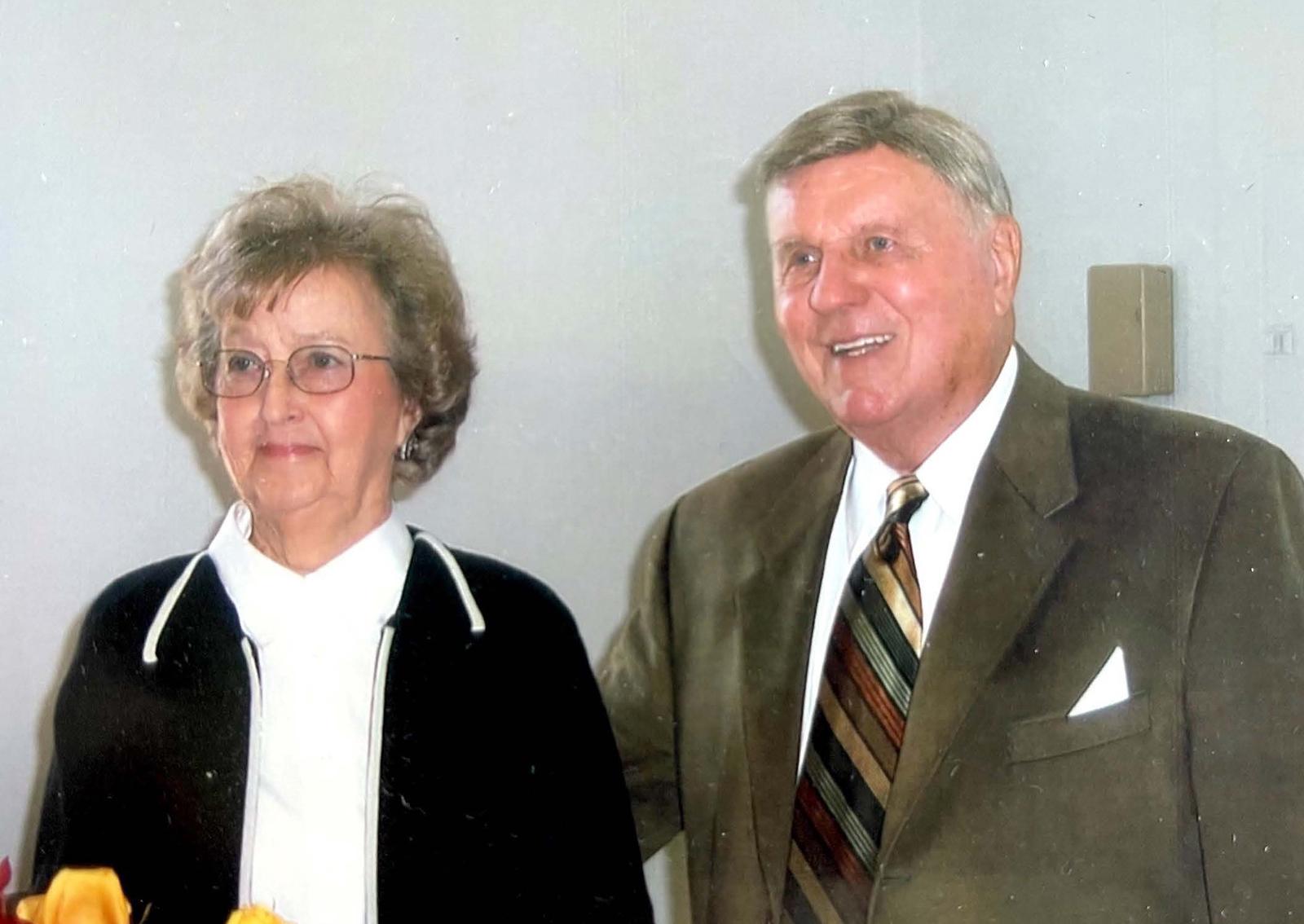 Older woman with glasses and short curly hair wearing black and white clothing stands next to an older man in a brown suit and striped tie smiling against a pla
