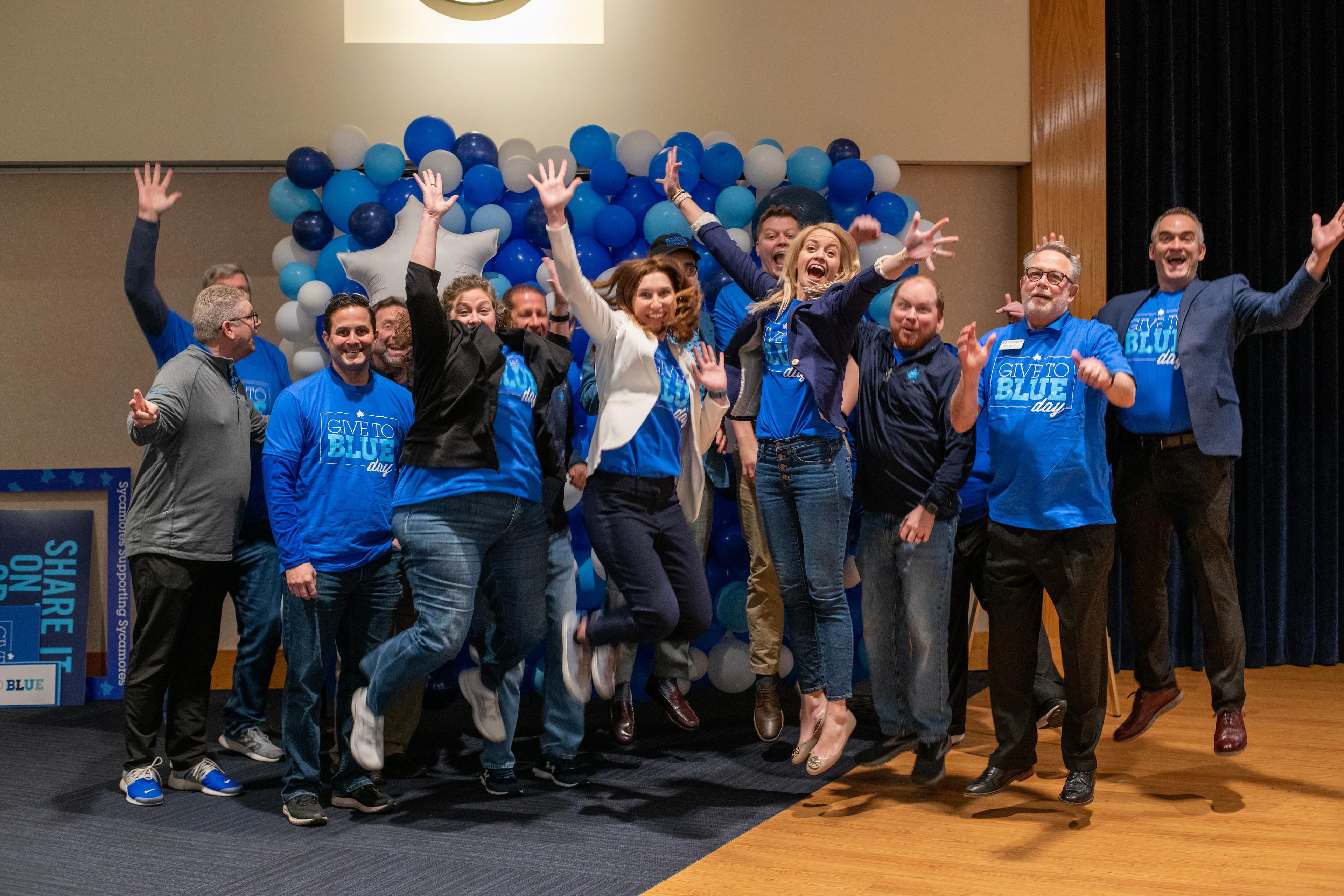 Group of people wearing blue Give to Blue Day shirts jumping and celebrating in front of blue and white balloon backdrop.