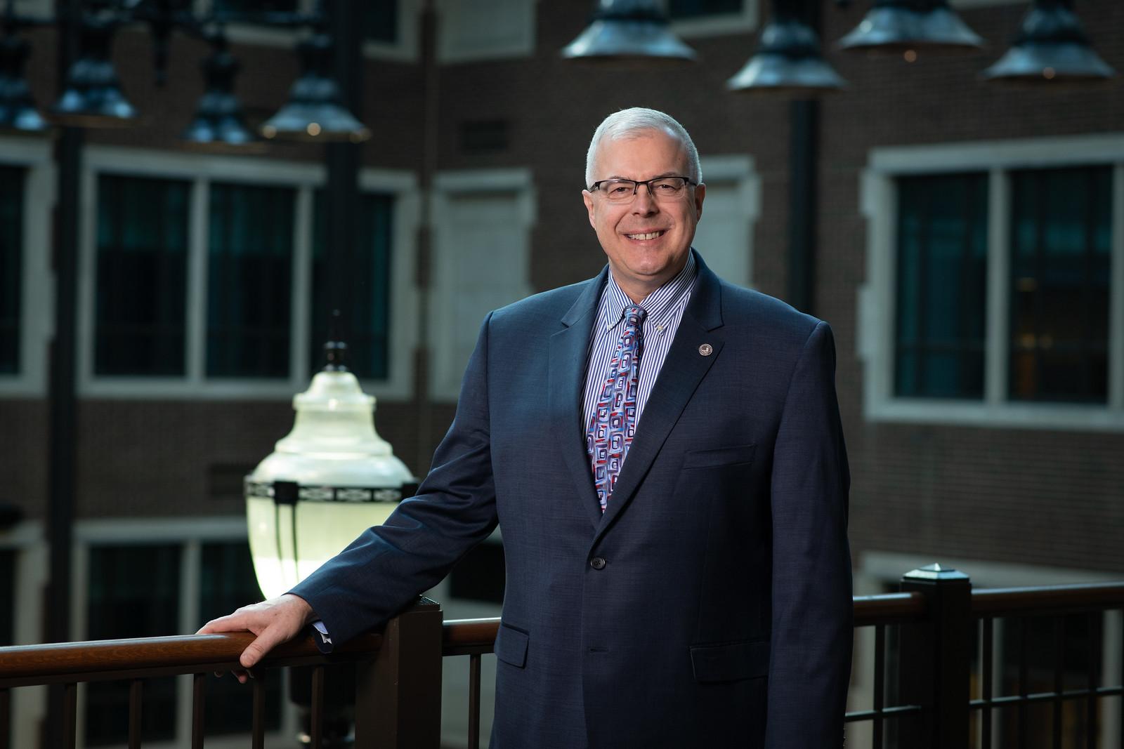 Middle-aged man with gray hair and glasses wearing a dark suit, patterned tie, and striped shirt, standing indoors by a wooden railing with brick walls behind.