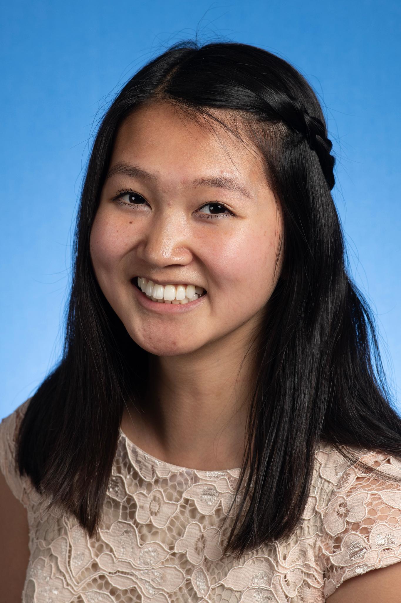 Young woman with long black hair braided on one side, smiling, wearing a light pink lace top against a blue background.