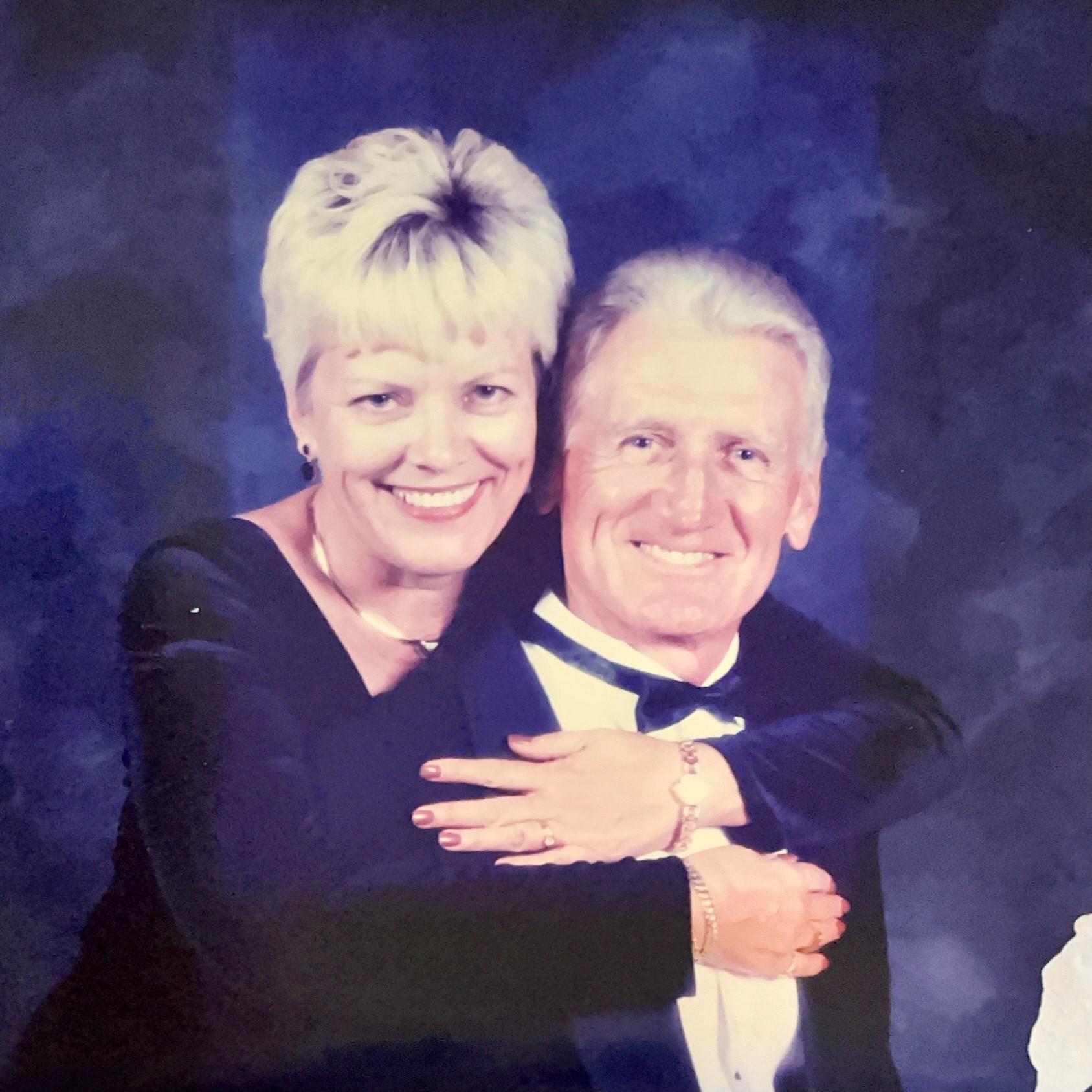 Smiling older couple dressed formally, woman with short blonde hair hugging man wearing a tuxedo, dark blue backdrop.