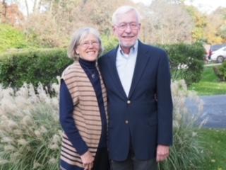 An older couple standing outdoors near greenery, smiling, wearing glasses, the woman in a striped vest and the man in a dark blazer.