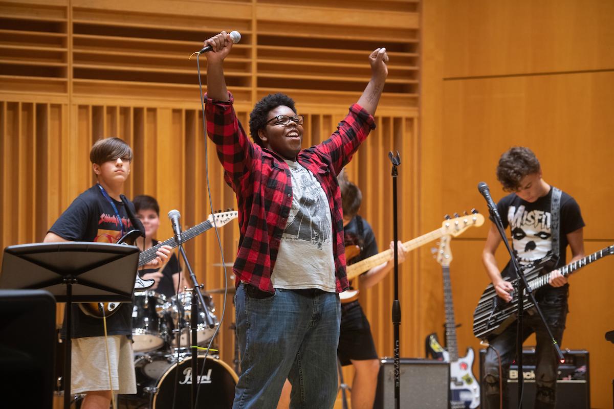 Teen musicians performing in a wood-paneled room, one singing with arms raised holding a microphone, others playing electric guitars and drums.
