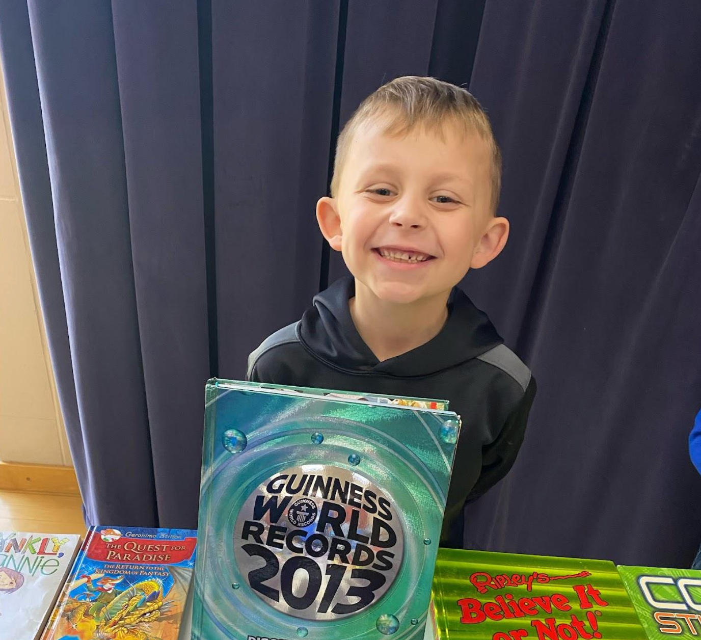 Young boy smiling while holding a Guinness World Records 2013 book, standing behind a table with colorful books displayed on it.