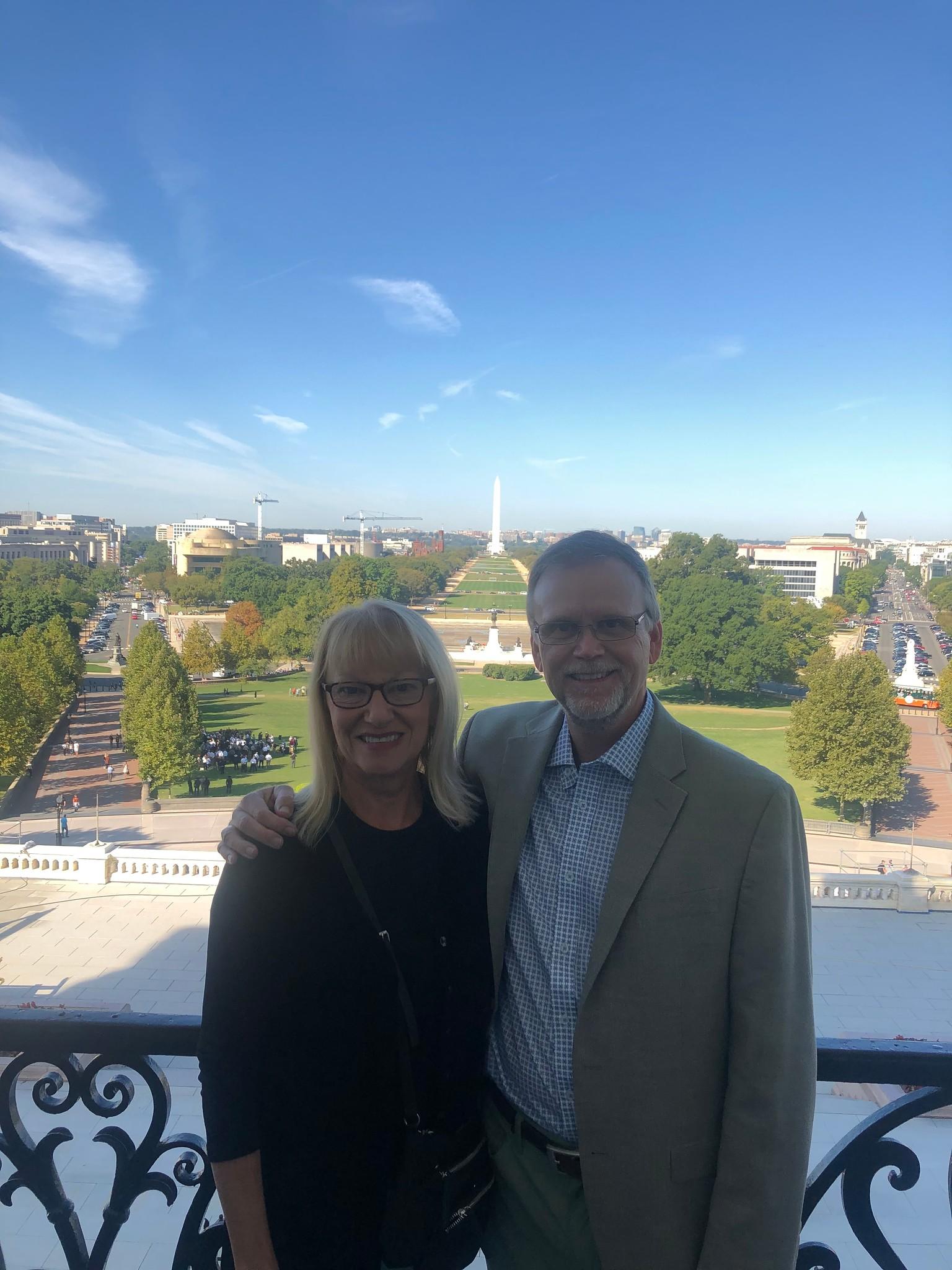 Smiling middle-aged couple with glasses standing close together on a balcony overlooking a green park and Washington Monument on a clear day.