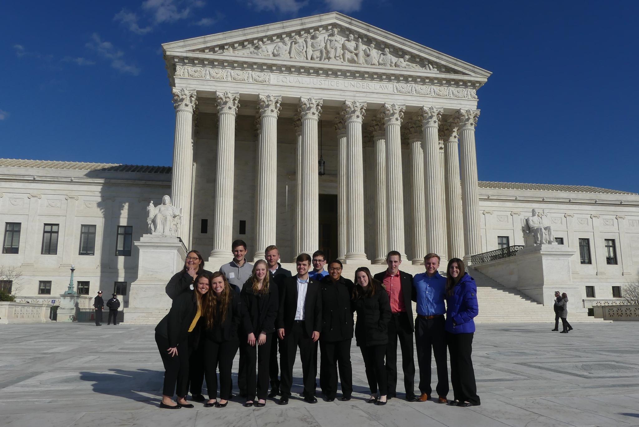 Group of 12 young adults standing in front of the Supreme Court building on a clear sunny day, dressed in business casual attire.