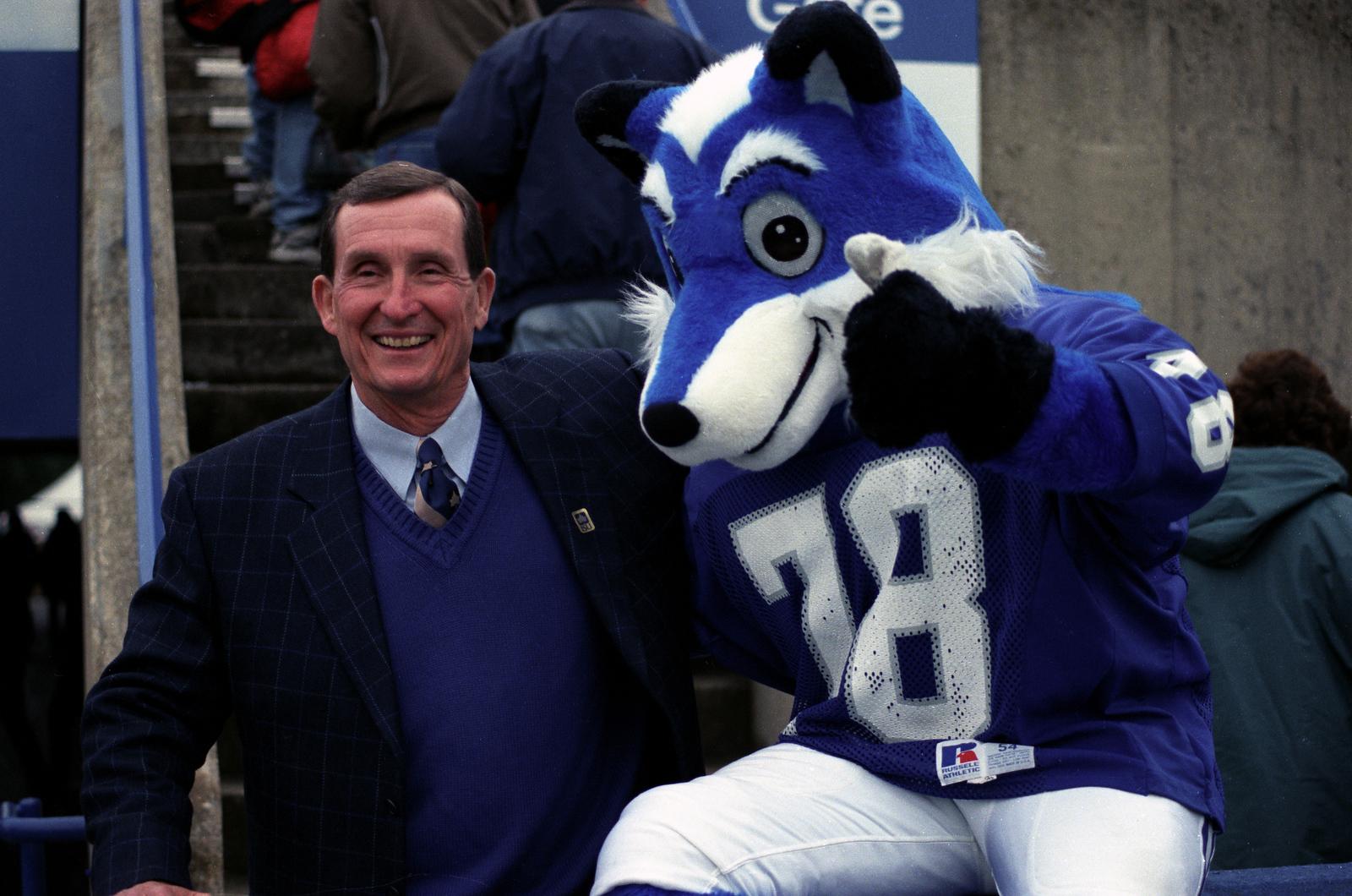 Smiling man in a suit and blue sweater poses with a person in a blue and white husky mascot costume wearing a number 78 jersey.