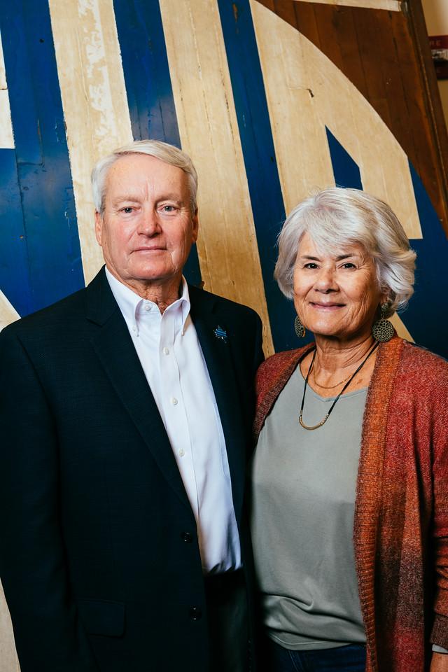 Older man in a dark suit and white shirt stands next to older woman in gray top and rust-colored cardigan against a blue and beige wall background.